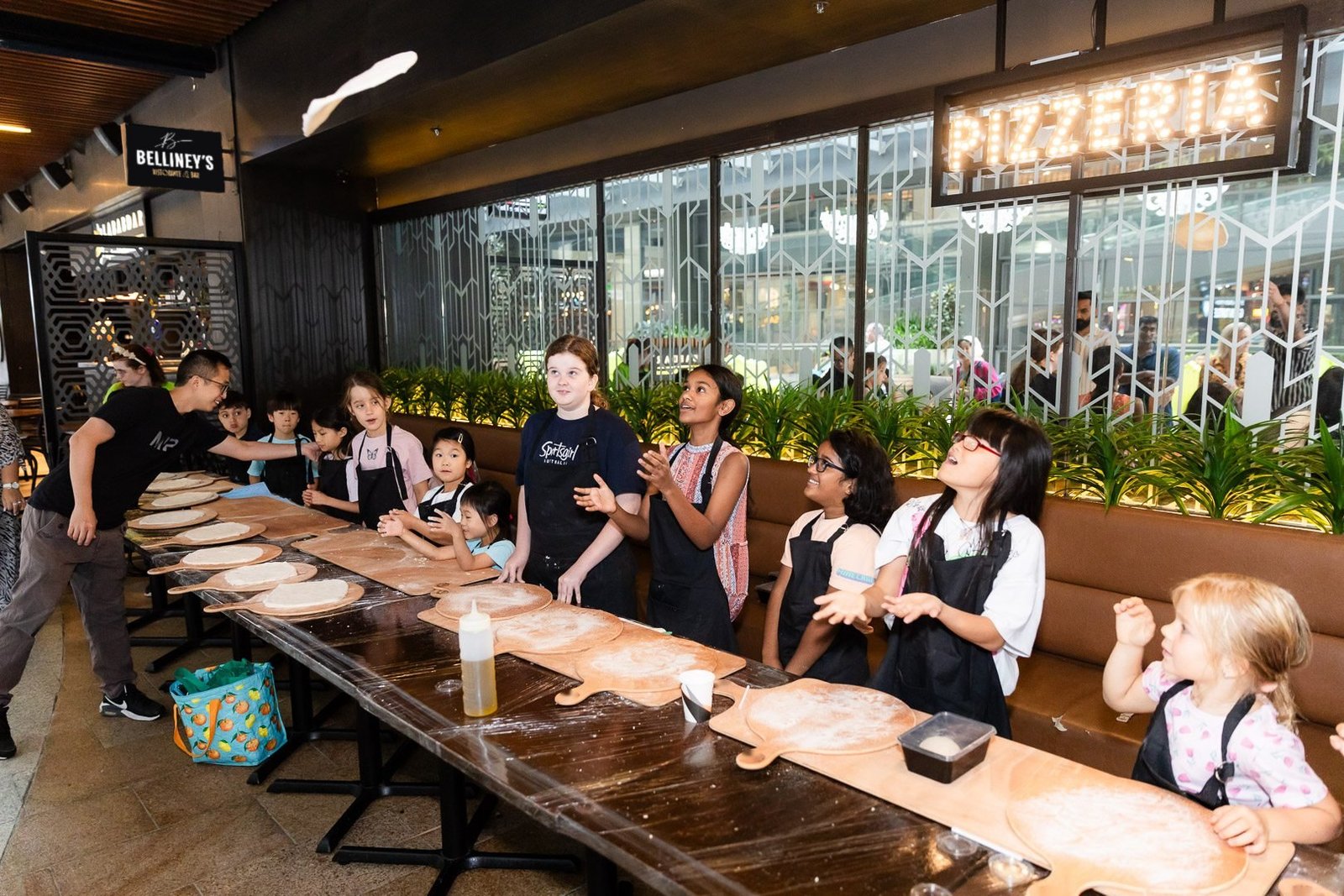 A row of kids in aprons watching a chef toss pizza dough